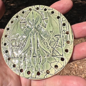 May include: A round, light green ceramic piece featuring a detailed relief of a Cthulhu-like creature with tentacles and wings. The design includes a cracked, textured background and small holes around the edge. The piece is held in a hand.