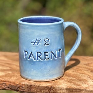 May include: A light blue ceramic mug with a glossy finish and a dark blue interior. The front of the mug features the embossed text #2 PARENT in dark blue lettering. The mug has a single curved handle on the right side and a cylindrical shape.