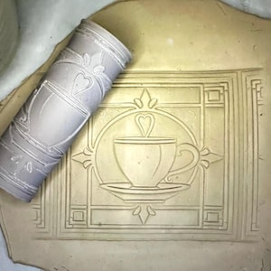 May include: A light grey cylindrical clay texture roller featuring a repeating embossed pattern of a coffee cup on a saucer with a heart-shaped steam swirl, shown pressing the design into a flat, beige, rectangular slab of soft, pliable clay.