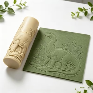 May include: A cylindrical beige rolling pin featuring a raised relief pattern of a long-necked dinosaur and foliage, paired with a square olive green flat stamp showing the same detailed dinosaur design in a pressed, embossed texture.