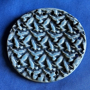 May include: A circular ceramic disc featuring a repeating pattern of embossed birds in flight. The surface has a glossy, light blue-grey finish with dark recessed areas between the bird shapes, creating a high-contrast, intricate, and textured design.