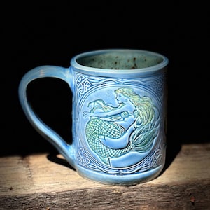 May include: A light blue ceramic mug with a handle, featuring a detailed relief of a mermaid. The mermaid has flowing blonde hair and a scaled tail. Celtic knotwork surrounds the mermaid. The mug is on a wooden surface.