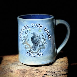 May include: Light blue ceramic mug with a dark blue interior and handle. The mug features a circular design with a raven perched on a branch, the words "SUPPORT YOUR LOCAL MURDER" are printed around the raven.