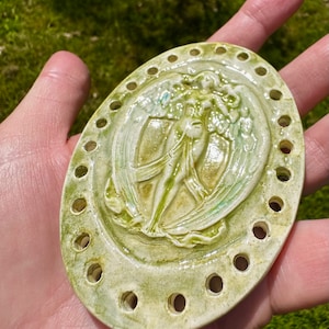 May include: A ceramic needle minder with a raised design of a winged figure in a flowing gown. The minder is oval shaped and has a white background with green and blue accents. There are 16 holes around the edge of the minder.