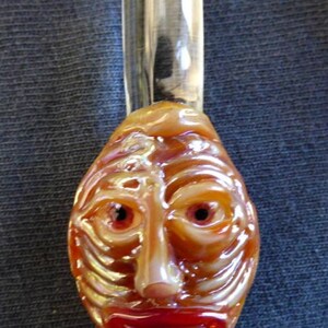 May include: A glass pendant featuring a brown face sculpted into the glass. The face has red eyes and a red mouth.