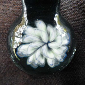 May include: A glass pendant with a white flower design with blue accents. The flower is encased in clear glass and the pendant is dark green.