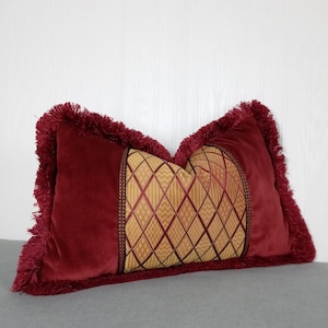 May include: A rectangular decorative pillow with a burgundy velvet exterior and a gold patterned center. The pillow has a fringe trim and is resting on a gray surface. The center features a diamond and stripe pattern.