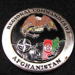 May include: A circular silver-toned metal challenge coin with a black enamel center. It features an eagle clutching an anchor, a NATO star emblem, and a map of Afghanistan colored with its national flag. The text reads REGIONAL COMMAND (SW) AFGHANISTAN.