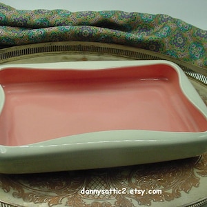 May include: A rectangular, vintage ceramic dish with a white exterior and a pink interior. The dish has a curved design and is displayed on a silver tray. The overall aesthetic is retro and mid-century modern.