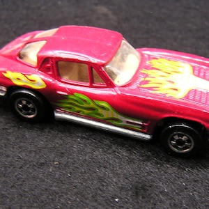 May include: A red toy car with a flame design in yellow and green. The car has black wheels and a silver trim. The car is a coupe style.