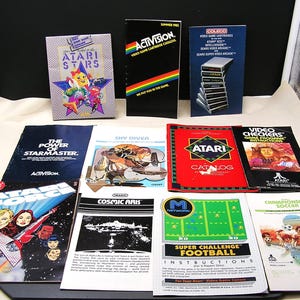 May include: A collection of vintage Atari video game manuals and catalogs featuring colorful graphic covers with titles including Atari Force, Cosmic Ark, Sky Diver, and Super Challenge Football, displayed in a grid on a light surface.