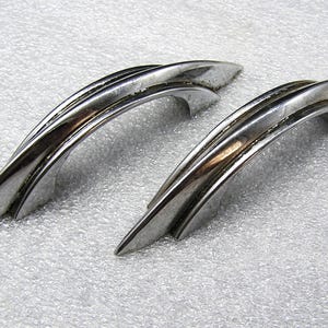 May include: Two matching silver-toned metal drawer pulls with a curved, arched design. Each handle features a sleek, elongated shape with pointed ends and a smooth, reflective finish, showcasing a distinct layered or tiered structural profile.