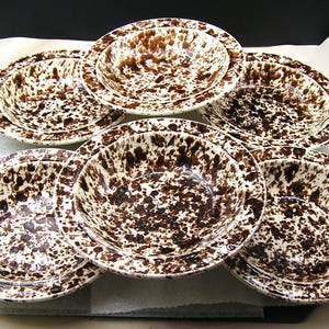 May include: A set of five round ceramic bowls featuring a speckled pattern with dark brown spots on a cream-colored background. Each bowl has a wide, flat rim and a deep, circular basin, arranged in a stacked formation to display the splatter design.