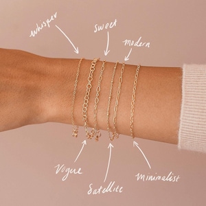 May include: A close-up of a woman's wrist wearing seven gold chain bracelets. The bracelets are labeled with names like "whisper", "sweet", "modern", "vogue", "satellite", and "minimalist".