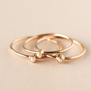 May include: Three delicate gold rings with small white pearl accents. The rings are stacked on top of each other.