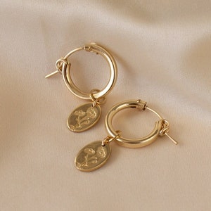 May include: A pair of gold-colored hoop earrings. Each earring has a small, oval charm with a floral design. The earrings are displayed on a beige satin surface, creating a soft, elegant aesthetic. The earrings are a classic jewelry item.