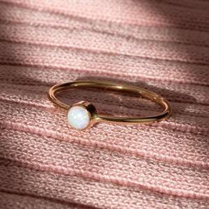 May include: A delicate gold ring with a white opal gemstone. The ring is on a pink knitted fabric background.