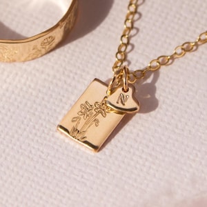May include: Two gold rings with a floral design and a gold necklace with a rectangular pendant featuring a floral design and a small heart charm with the letter "N" engraved on it.
