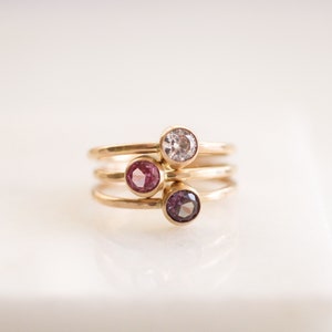 May include: A set of three gold rings with small gemstones. The rings are stacked on top of each other. The gemstones are a light pink, a dark pink, and a clear white.