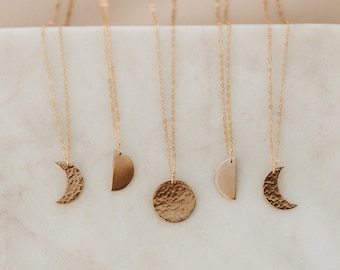 Può includere: Cinque collane d'oro con diversi ciondoli a forma di luna e mezzaluna. Le collane sono su una superficie bianca.