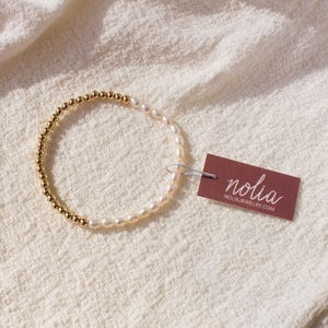 May include: A gold and white beaded bracelet with a tag that reads "nolia" and "nolijajewelry.com".