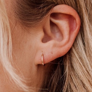 May include: A close-up of a person's ear with a small gold hoop earring. The earring is simple and delicate, and it is a popular choice for everyday wear.