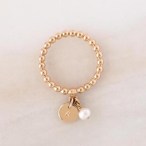 May include: A gold beaded ring with a charm featuring the letter "R" and a small pearl.