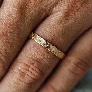 May include: A thin gold band ring with a textured surface. The ring is worn on a finger.