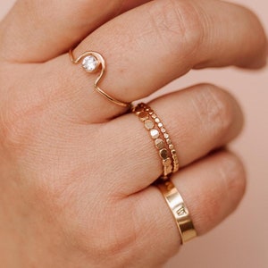 May include: A hand wearing several gold rings. One ring features a small, clear gemstone. Another ring has a beaded design, and a third ring is a simple band. The rings are set against a soft pink background.