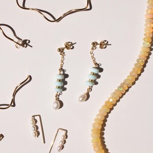May include: Gold-toned jewelry assortment on a white background. Includes dangling earrings with light blue beads and a pearl, a necklace of small, light-colored stones, and other earrings with pearls.