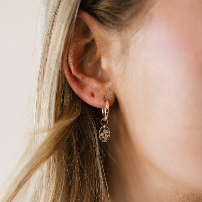 May include: Gold hoop earrings with a small oval pendant. The pendant features a floral design. The earrings are shown on a person's ear, with blonde hair visible in the background. The earrings are a simple and elegant accessory.