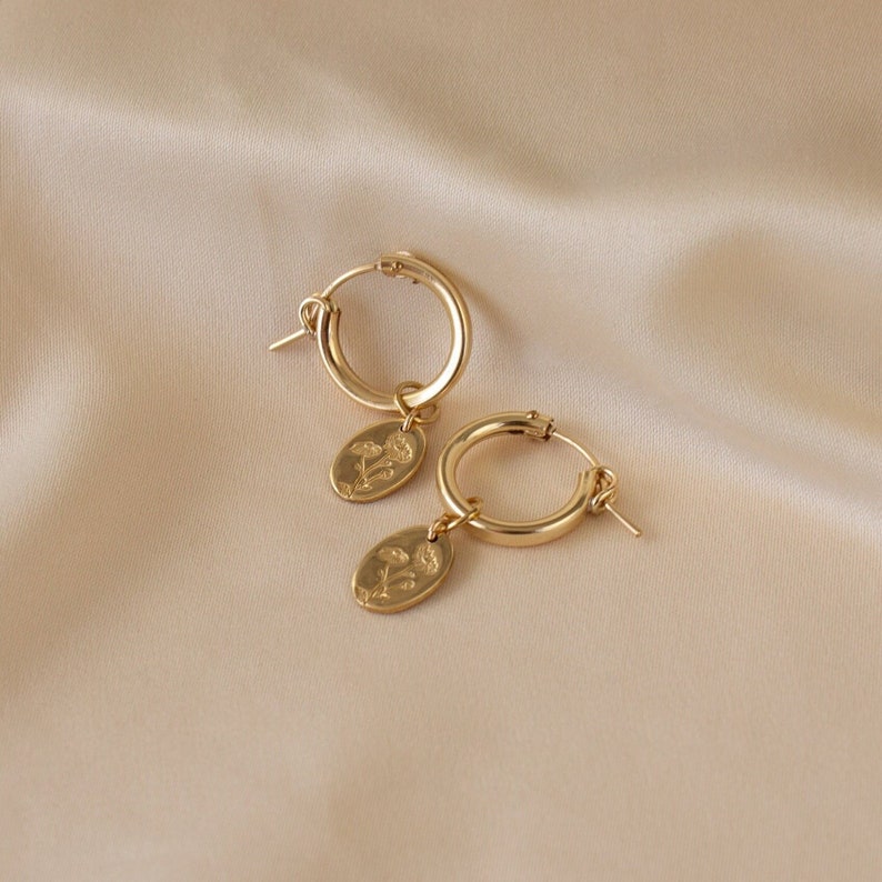 May include: A pair of gold-colored hoop earrings. Each earring has a small, oval charm with a floral design. The earrings are displayed on a beige satin surface, creating a soft, elegant aesthetic. The earrings are a classic jewelry item.