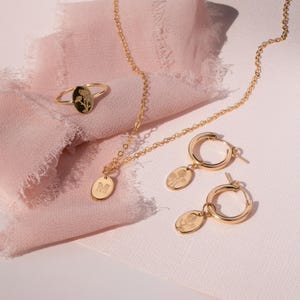 May include: A gold-toned jewelry set featuring a ring, necklace, and hoop earrings. Each piece has a floral design on an oval pendant. The necklace has an initial "M" pendant. The jewelry is displayed on a pink fabric.