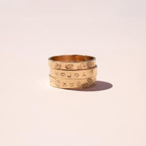 May include: A stack of three gold-toned metal rings. The top ring features an engraved floral pattern. The middle ring is stamped with the letters W, J, and A separated by heart symbols. The bottom ring displays the letters M and S with a heart.