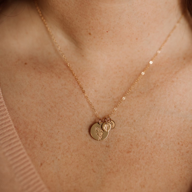 May include: A gold chain necklace with three charms. The largest charm features a floral design, while the other two charms are smaller and have a simple design.