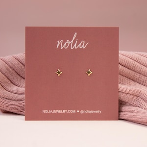 May include: A pair of gold star-shaped stud earrings on a pink card with the word "nolia" printed in white. The card also includes the website "nolijajewelry.com" and the social media handle "@nolijajewelry".