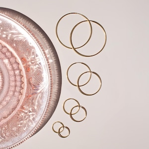 May include: A collection of gold-colored hoop earrings of varying sizes, displayed against a soft pink background. The earrings are arranged in pairs, with a decorative pink plate in the upper left corner.
