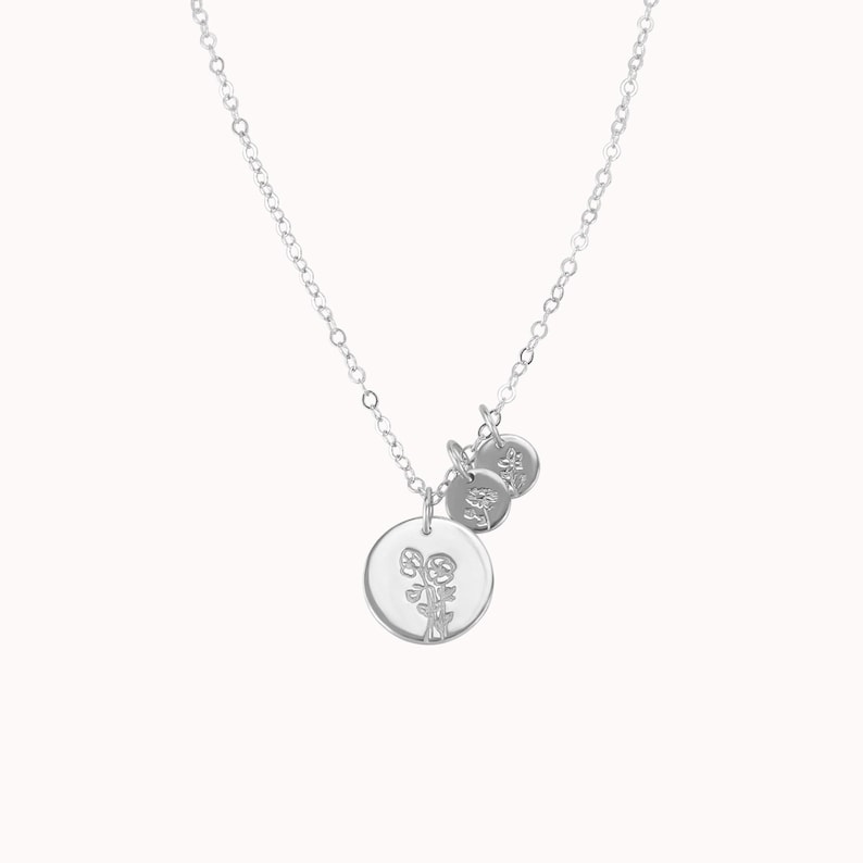 May include: A silver necklace with a delicate chain and three charms. The largest charm is a circle with a floral design. The two smaller charms are circles with a simple design.