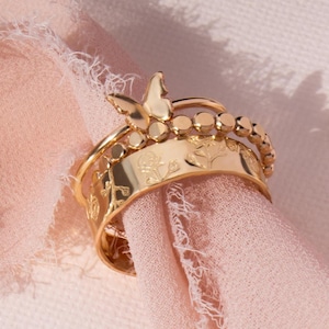 May include: Gold-toned ring set with a delicate butterfly charm and floral engravings. The rings are stacked on top of each other, creating a layered look.