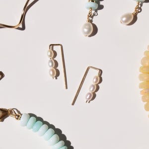 May include: Gold-tone earrings with three small, off-white pearls. The earrings have a curved design, with the pearls clustered together. Other jewelry pieces, including a bracelet with blue beads and other earrings, are also visible.