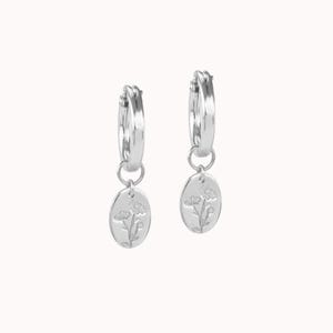 May include: A pair of silver hoop earrings with oval pendants. Each pendant features a floral design. The earrings are displayed against a white background, highlighting their polished, reflective surface. The earrings are a jewelry item.