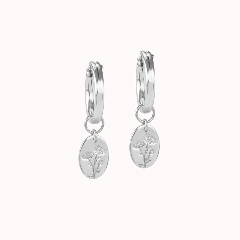 May include: A pair of silver hoop earrings with oval pendants. Each pendant features a floral design. The earrings are displayed against a white background, highlighting their polished, reflective surface. The earrings are a jewelry item.