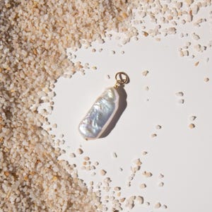 May include: A lustrous, elongated pearl pendant with a gold-colored bail. The pearl has a pearlescent sheen and a unique, irregular shape. The pendant is set against a backdrop of small, light-colored pebbles and a white surface.