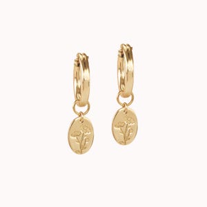 May include: A pair of gold-colored hoop earrings. Each earring has a small, oval pendant with a floral design. The earrings are displayed against a plain, light background.