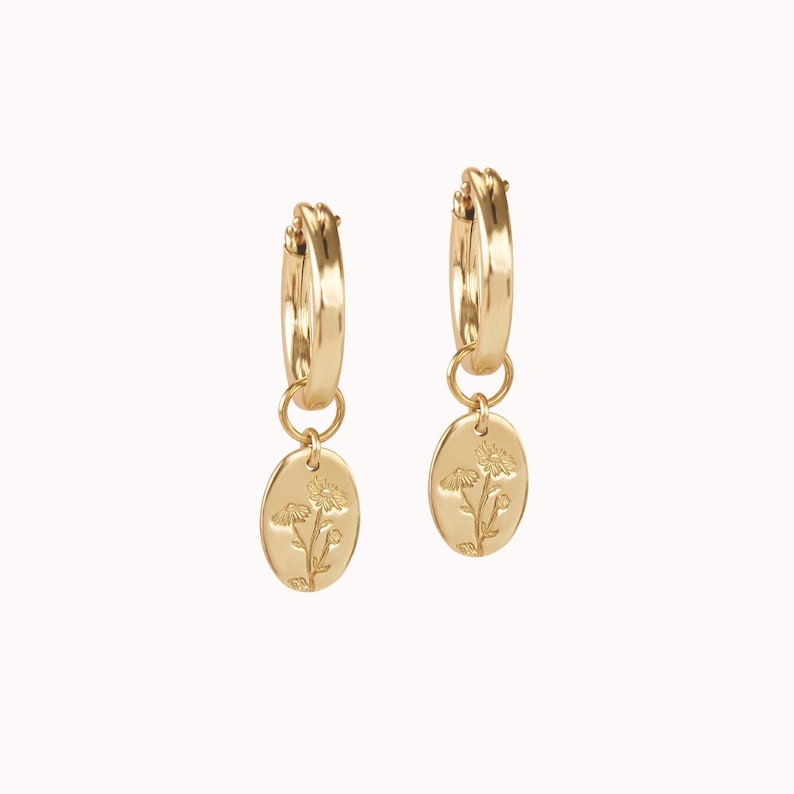 May include: A pair of gold-colored hoop earrings. Each earring has a small, oval pendant with a floral design. The earrings are displayed against a plain, light background.