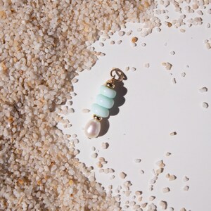 May include: A gold-colored charm with a light blue stone stack and a pearl. The charm is on a white surface with sand-like granules. The charm has a small loop at the top for attachment.