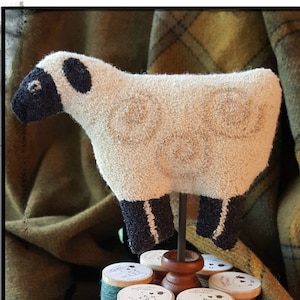 Op de afbeelding: Een houten spoelhouder met zes spoelen garen in verschillende tinten groen en bruin. De houder is getooid met een wit en zwart wollen schaapje. De tekst "The Woolly Thread Co." is zichtbaar op de basis van de houder.