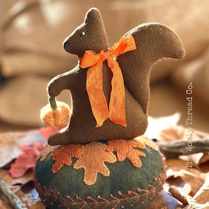 May include: A brown felt squirrel with an orange ribbon holding a small orange felt pumpkin. The squirrel is standing on a green felt pincushion with embroidered brown and orange leaves. The pincushion is surrounded by brown leaves.