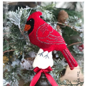 Könnte beinhalten: Ein roter Kardinal mit grauen Flügeln sitzt auf einer weißen, flauschigen Basis auf einer alten Metallglocke mit einem roten Band. Die Glocke steht auf zwei übereinander gestapelten, mit Moos bedeckten Ringen. Der Hintergrund ist ein schneebedeckter Nadelbaum.