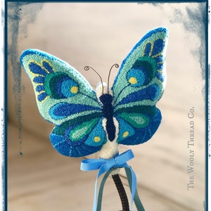 May include: A blue and green butterfly with yellow accents is perched on a vintage oil can. The butterfly is made of yarn and has a 3D effect. The oil can is rusty and has a blue ribbon tied around it.